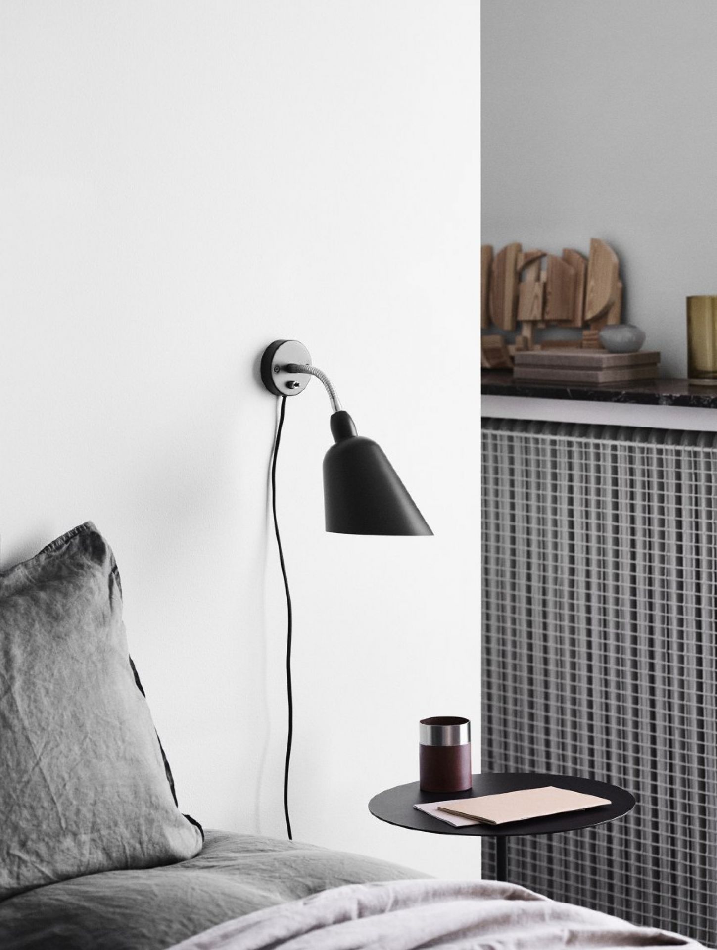 Schwarze Bellevue Wandleuchte von AndTradition neben einem Bett mit grauem Kissen und Nachttisch.