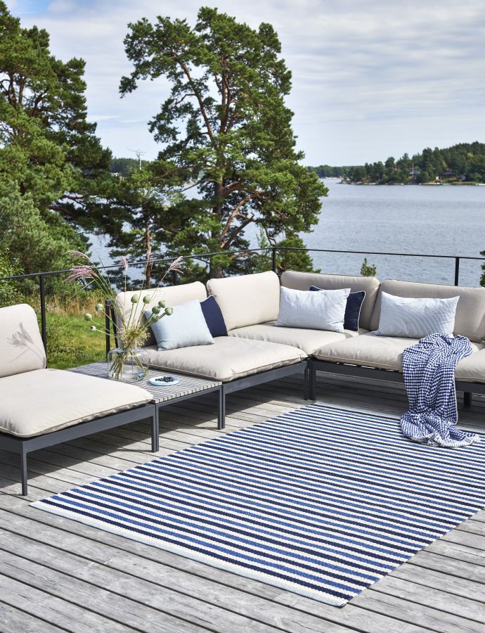 Teo Teppich Denim von Pappelina, gestreifter Outdoor Teppich in Blau-Weiß auf einer Holzterrasse mit Lounge-Möbeln.