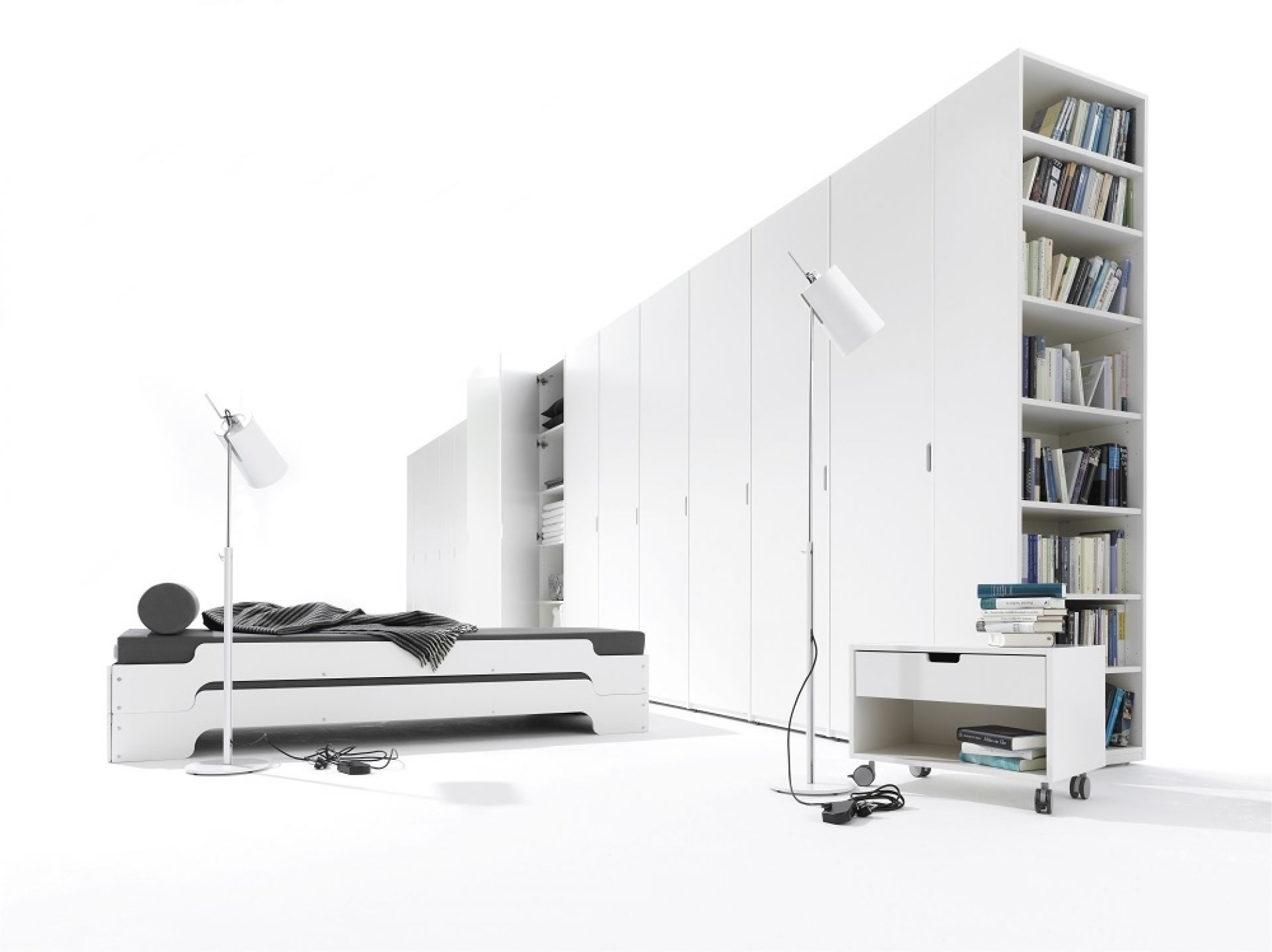 Weißer Müller Möbelwerkstätten Nachttisch mit Schublade und Rollen vor weißem Kleiderschrank und Bücherregal.