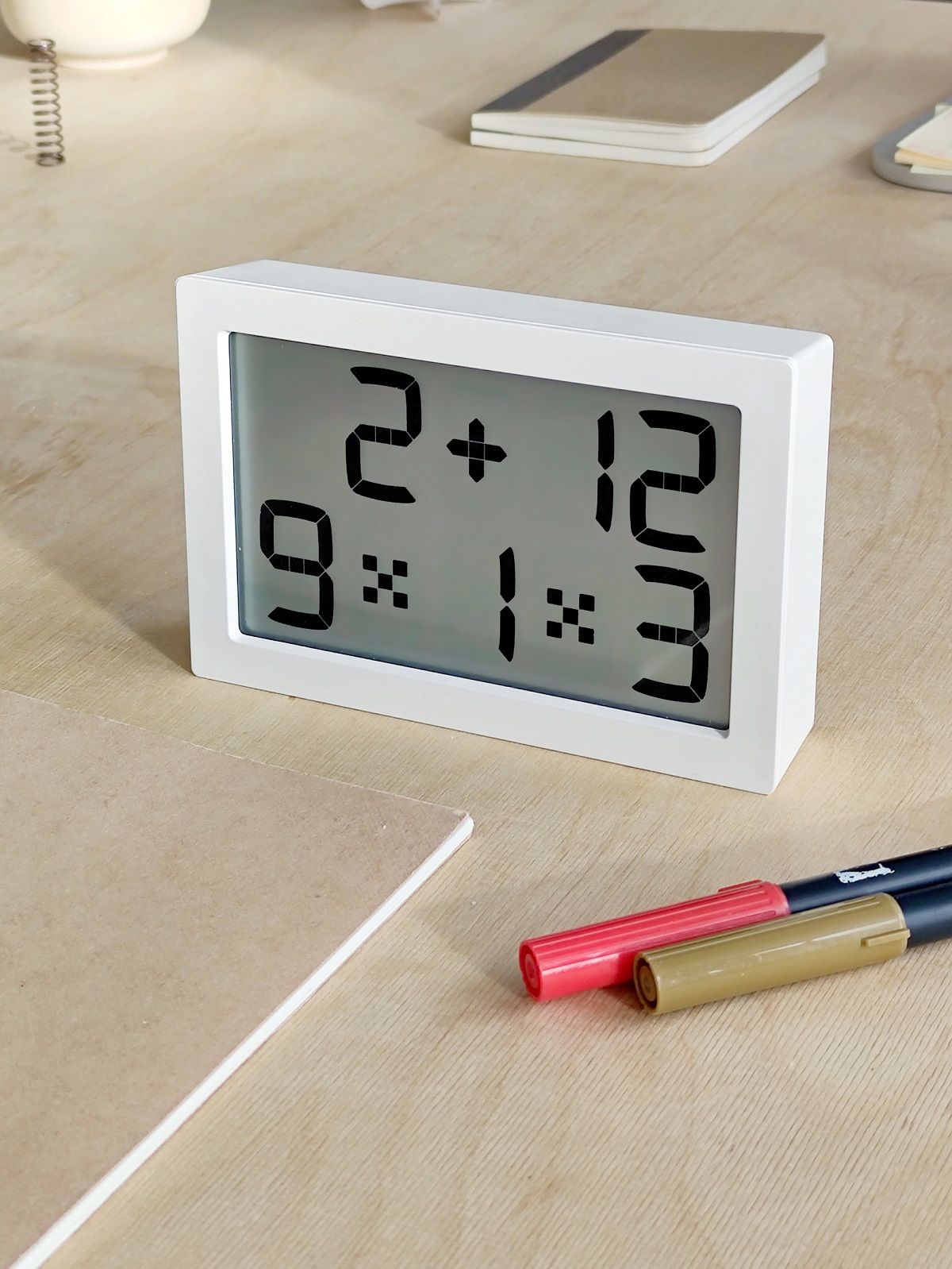 Weiße Digitaluhr (Albert Clock) auf einem Schreibtisch mit Stiften und Notizbüchern. Tischuhr, Wanduhr.