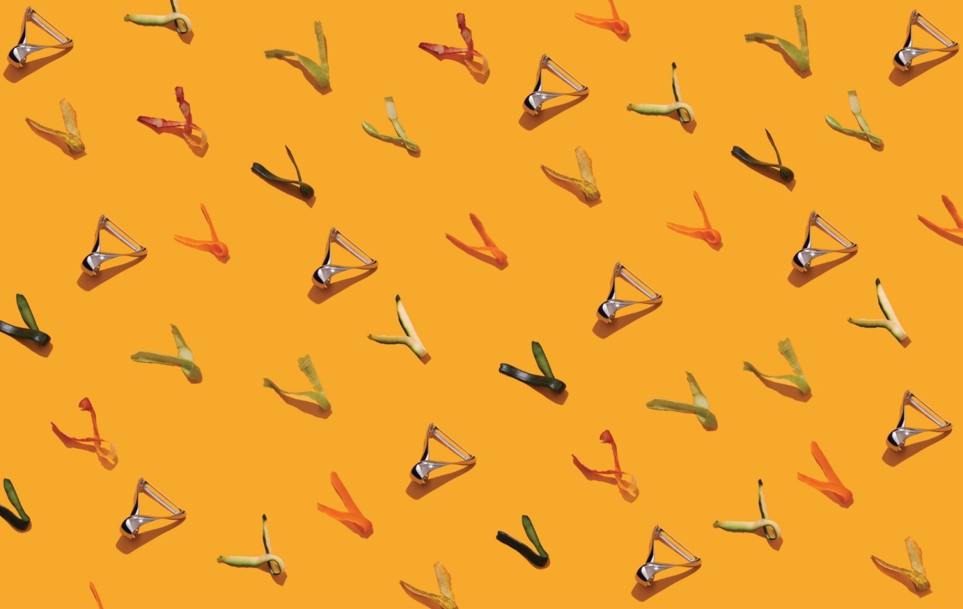 Musterbild: Alessi Sfrido Kartoffelschäler aus Edelstahl mit bunten Gemüseschalen auf orangefarbenem Hintergrund.