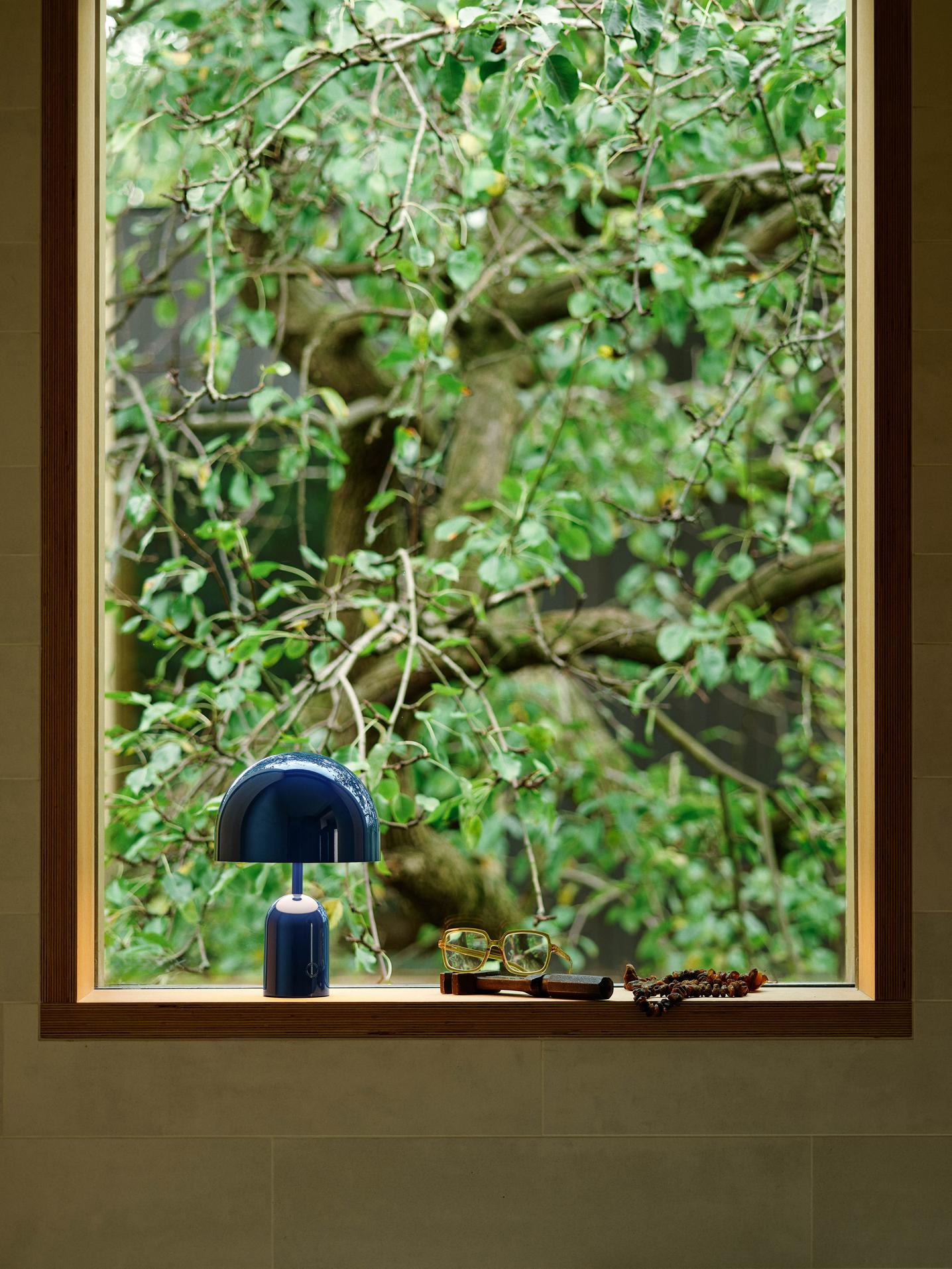 Blaue Bell LED Akkuleuchte von Tom Dixon auf Fensterbank mit Baum im Hintergrund.