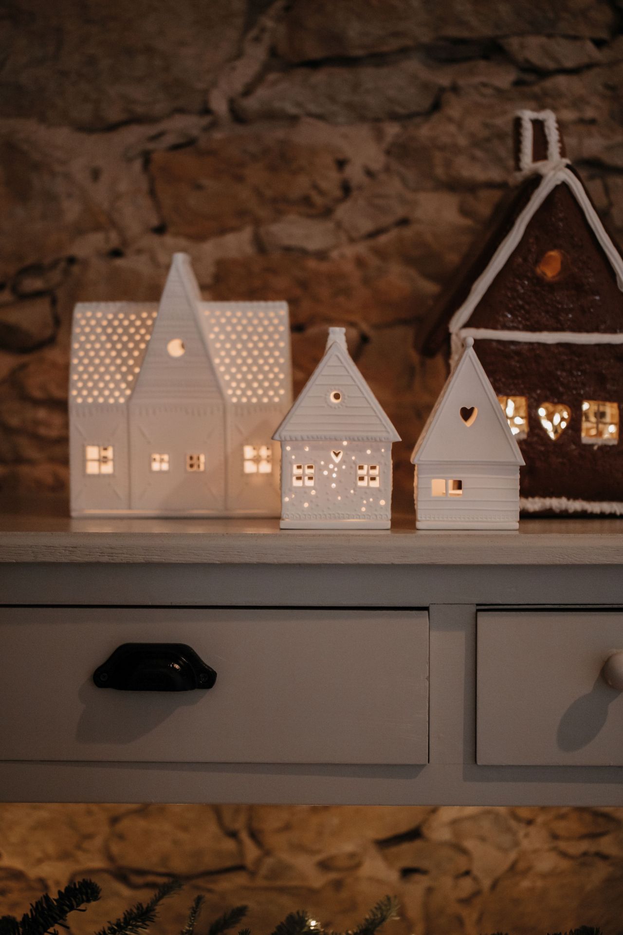 Drei beleuchtete Lebkuchenhäuser, darunter ein Räder Lichthaus, auf einer Kommode vor einer Steinwand.