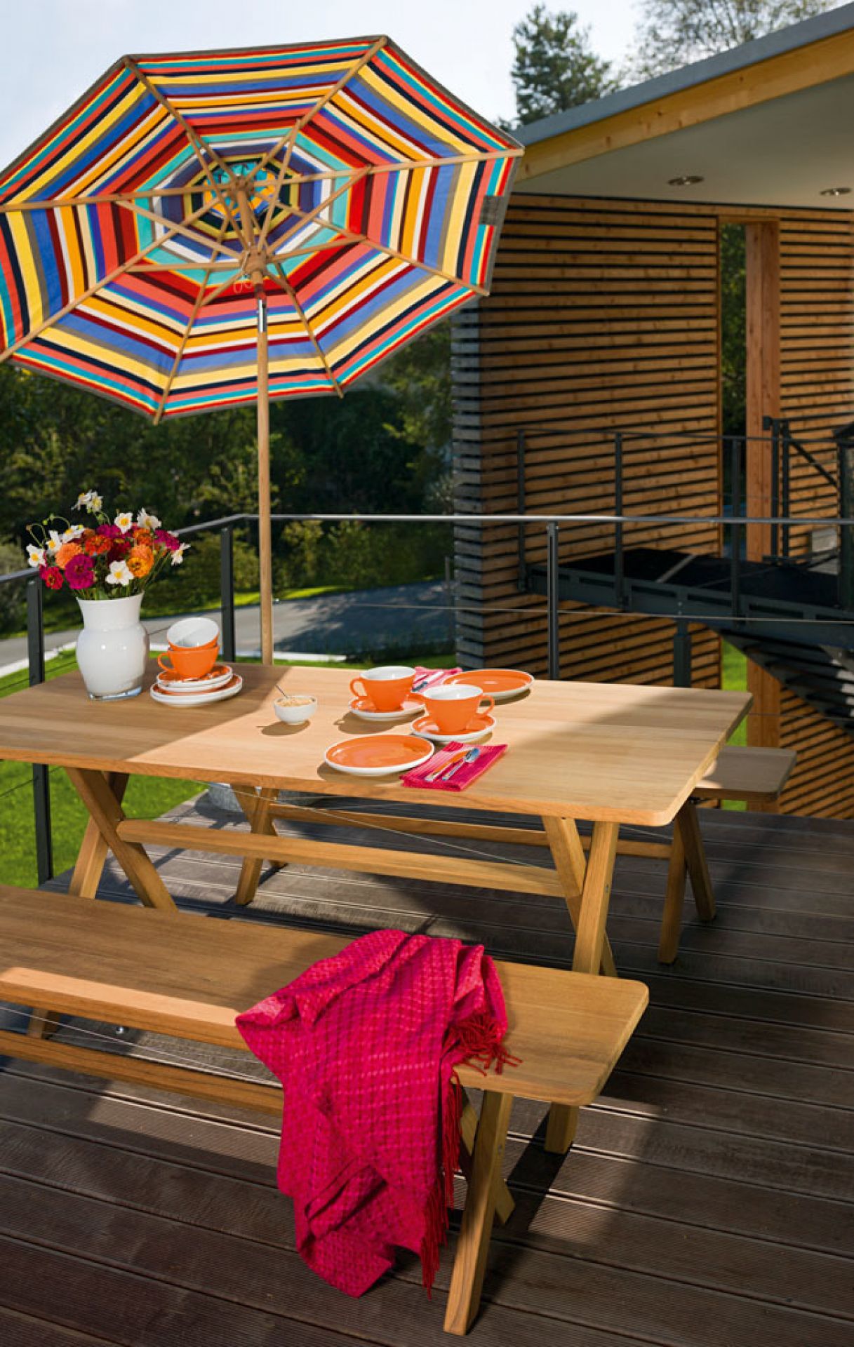 Weishäupl Cross Tisch klappbar aus Holz mit Bänken und farbenfrohem Sonnenschirm auf Terrasse.