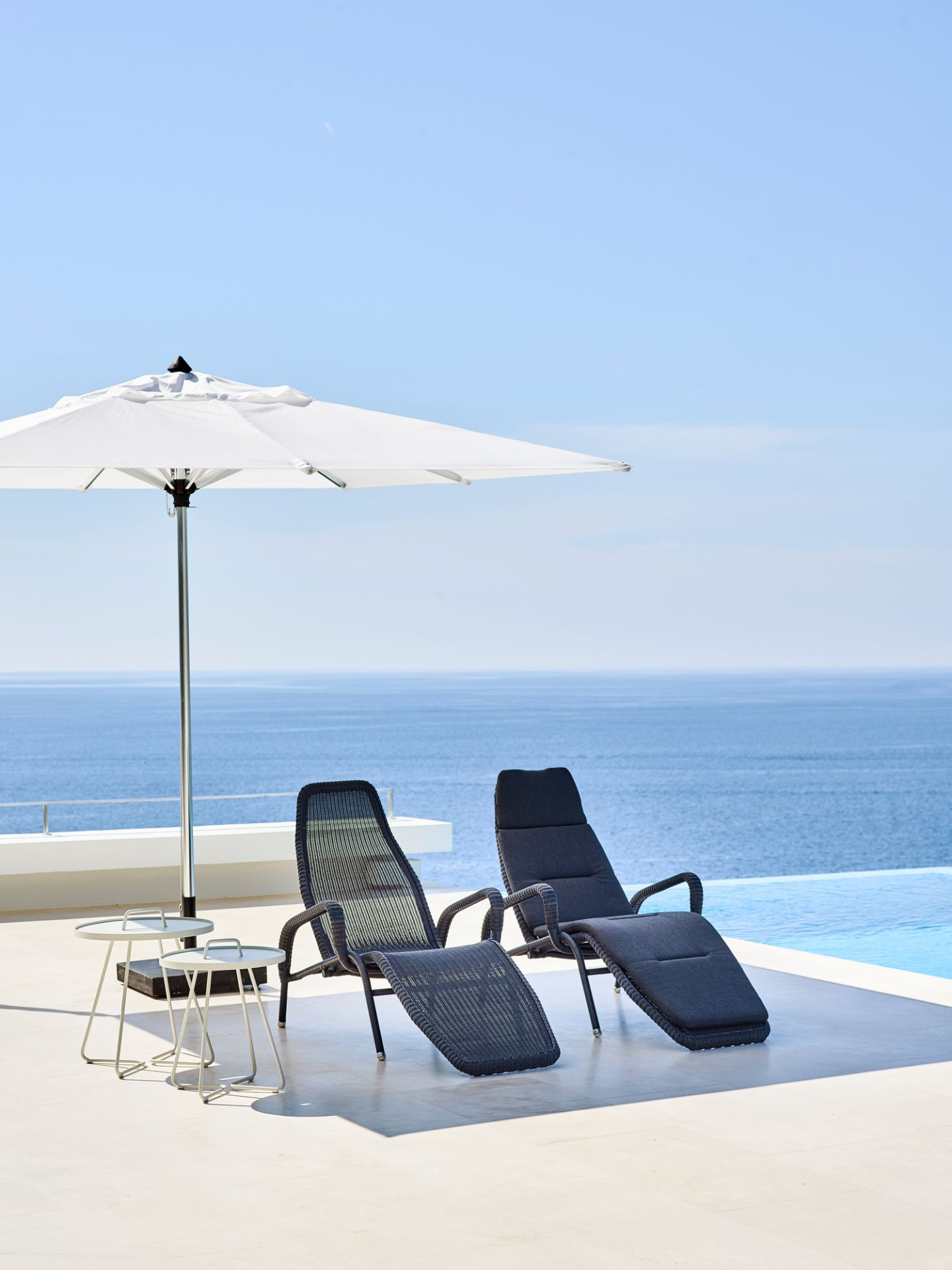 Zwei Cane-Line Sonnenliegen mit Sonnenschirm und Beistelltischen am Pool mit Meerblick.