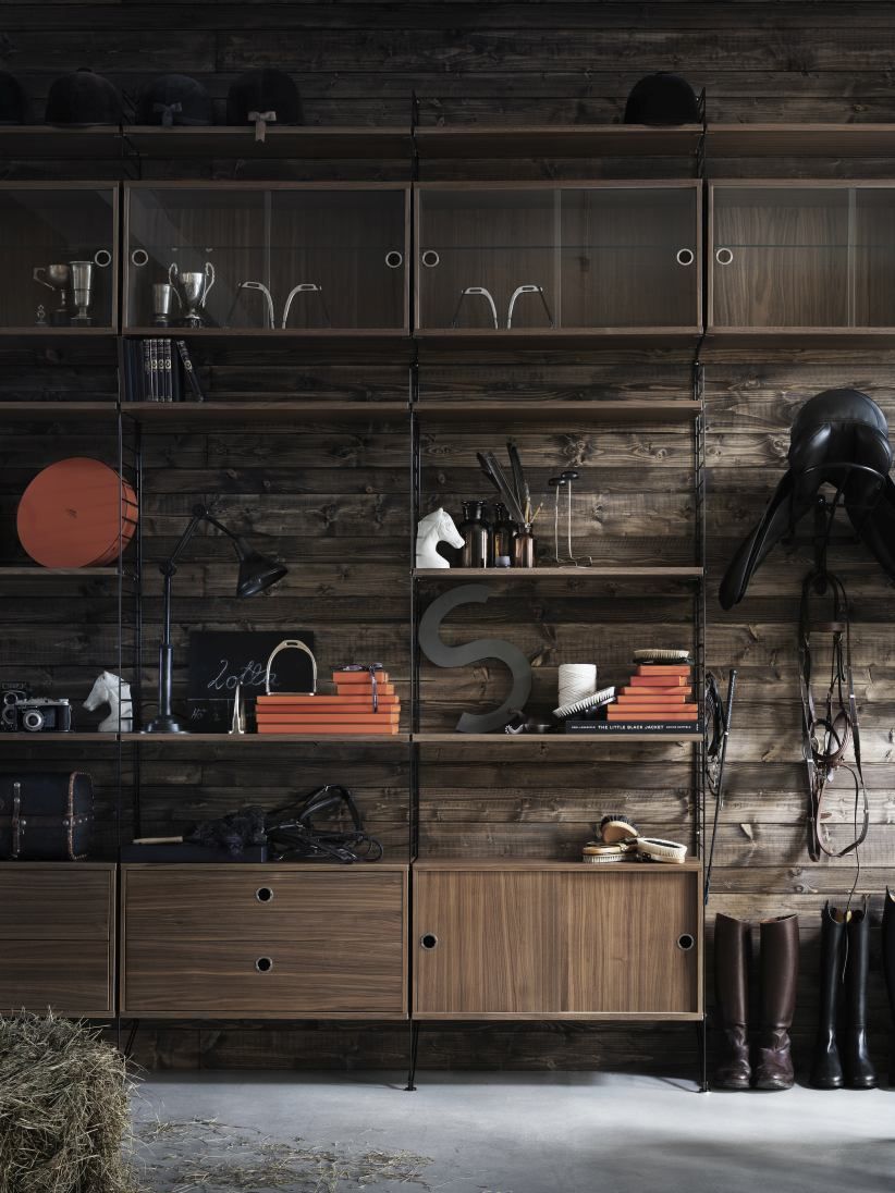 Wohnzimmer KONFIGURATIONEN L8 String Furniture Regalsystem aus Holz mit Reitsport-Accessoires vor rustikaler Holzwand.