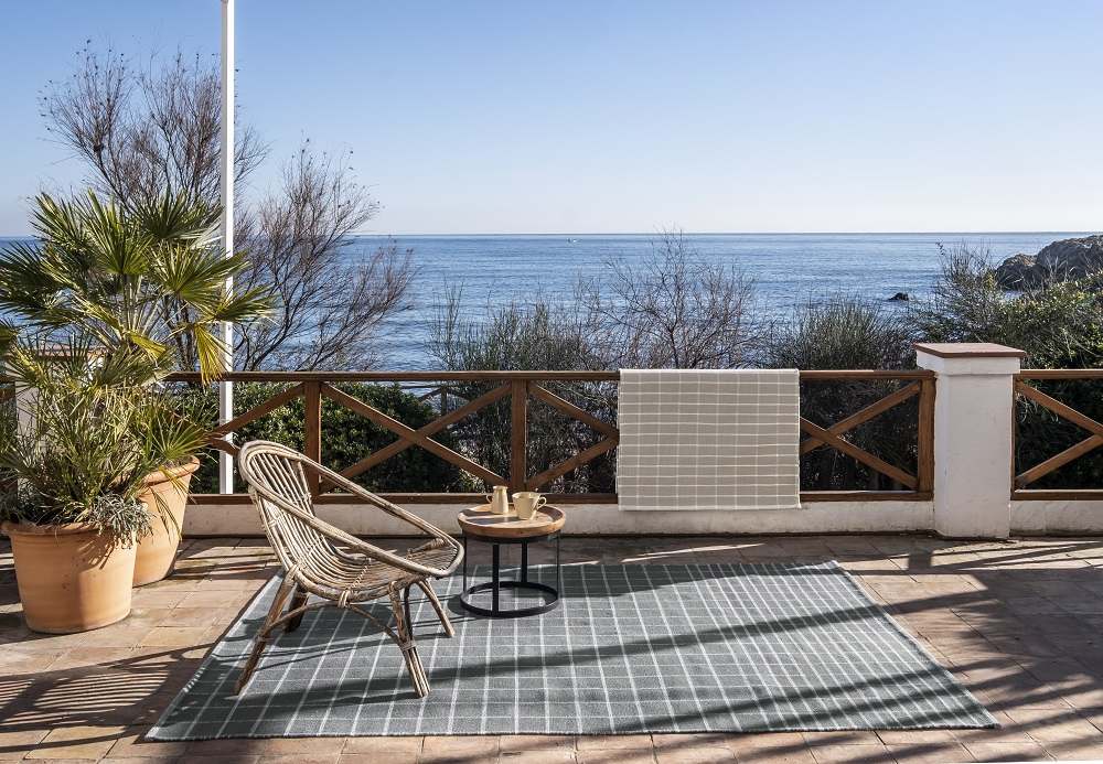 Nanimarquina Tiles Collection Teppich #3, 170x240 cm, auf einer Terrasse mit Meerblick, mit Stuhl und Beistelltisch.