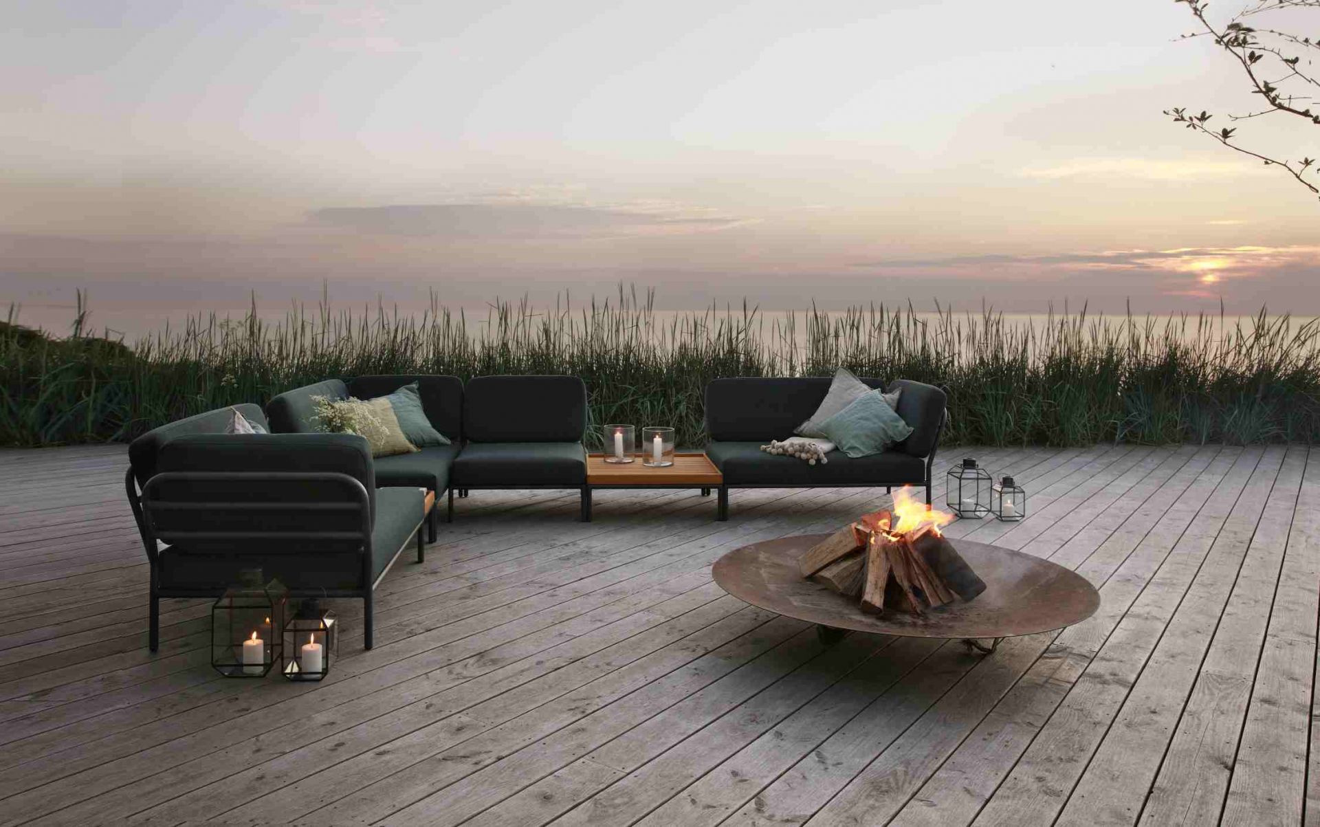 Houe Level Outdoor Couchtisch mit Lounge-Möbeln und Feuerstelle auf einer Holzterrasse bei Sonnenuntergang.