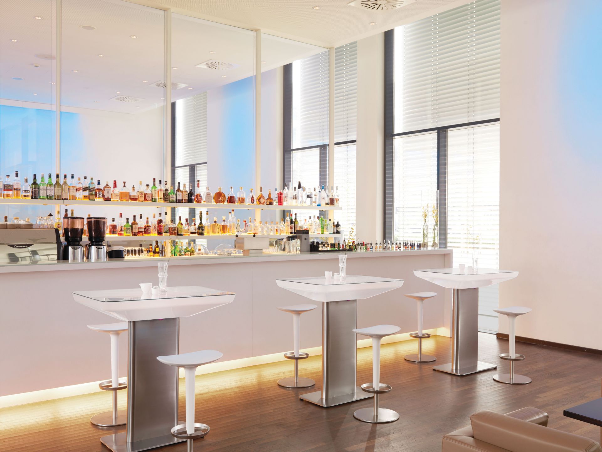 Heller Raum mit Moree Stehtischen und Barhockern vor einer Bar mit Spirituosen.