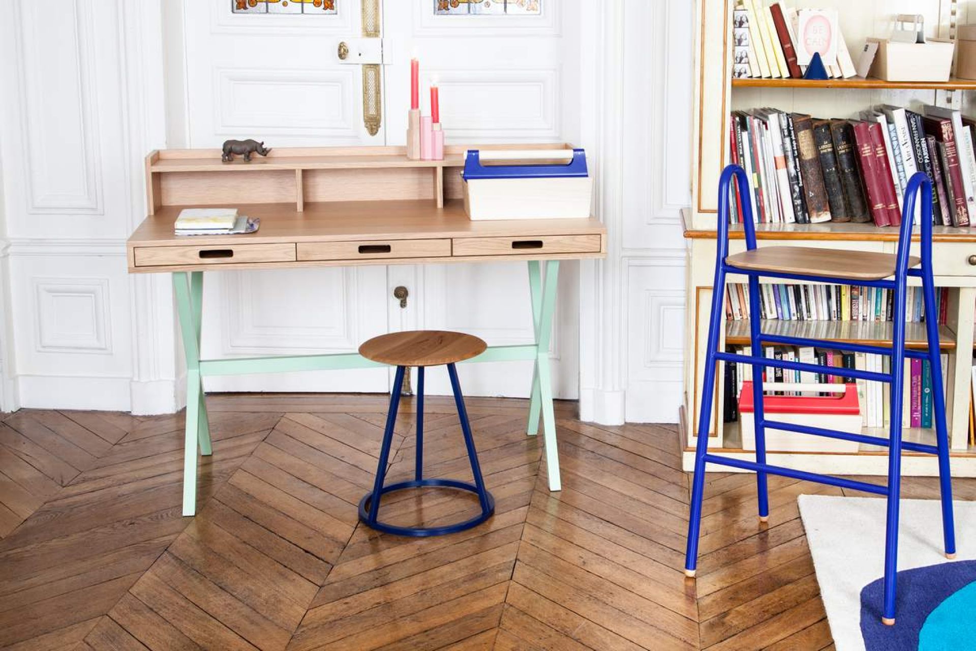 Gustave Hocker von Hartô, runder Sitzhocker mit blauem Metallgestell vor Schreibtisch und Bücherregal.