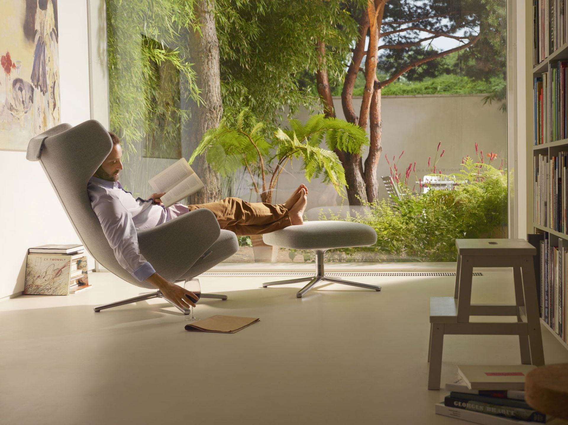 Mann entspannt im grauen Vitra Sessel mit Hocker, liest Buch, modernes Wohnzimmer.