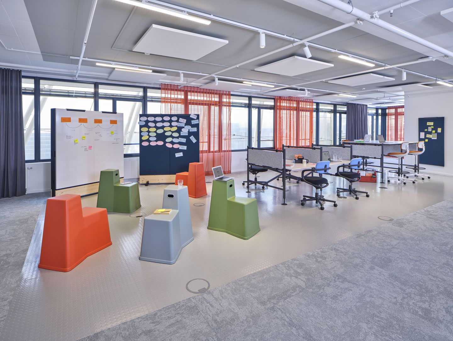 Großraumbüro mit Vitra Rookie High Bürodrehstühlen an höhenverstellbaren Schreibtischen, ergänzt durch farbenfrohe Sitzhocker für flexible Arbeitsbereiche.