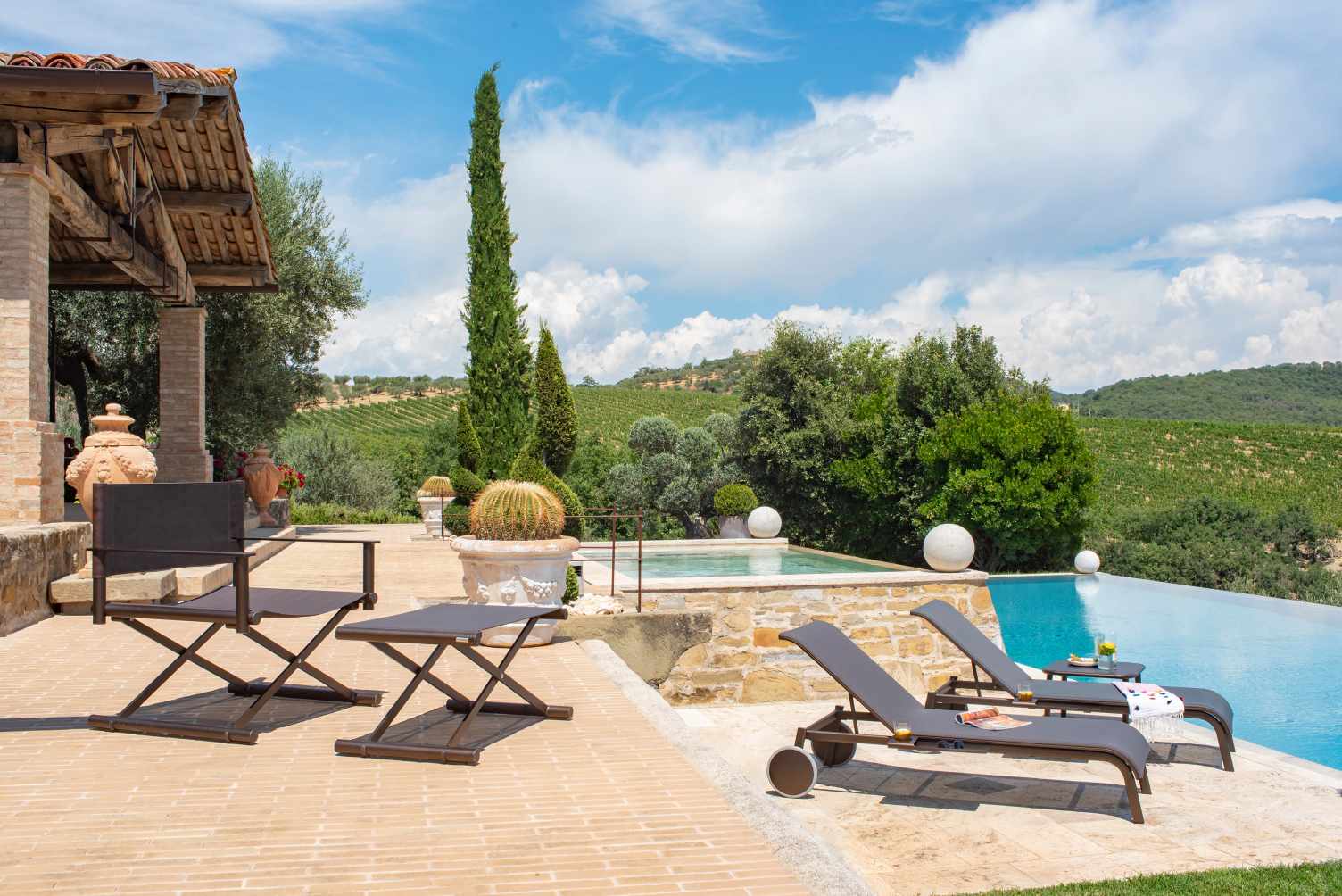 Terra Fußhocker von Emu, Outdoor-Möbel aus Metall, braun, vor Pool und toskanischer Landschaft.