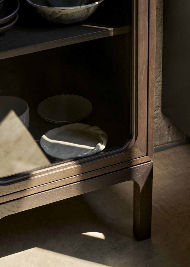 Detailaufnahme des AndTradition Trace SC87 Schranks aus dunklem Holz mit Glasfront und Geschirr.