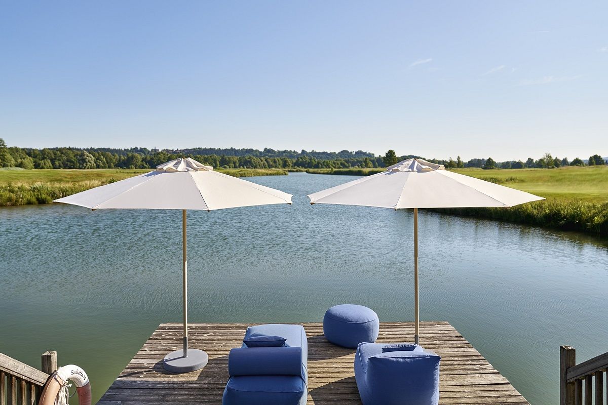 Zwei weiße Weishäupl Sonnenschirme Ø 335 cm auf einem Holzsteg am See mit blauen Sitzmöbeln.