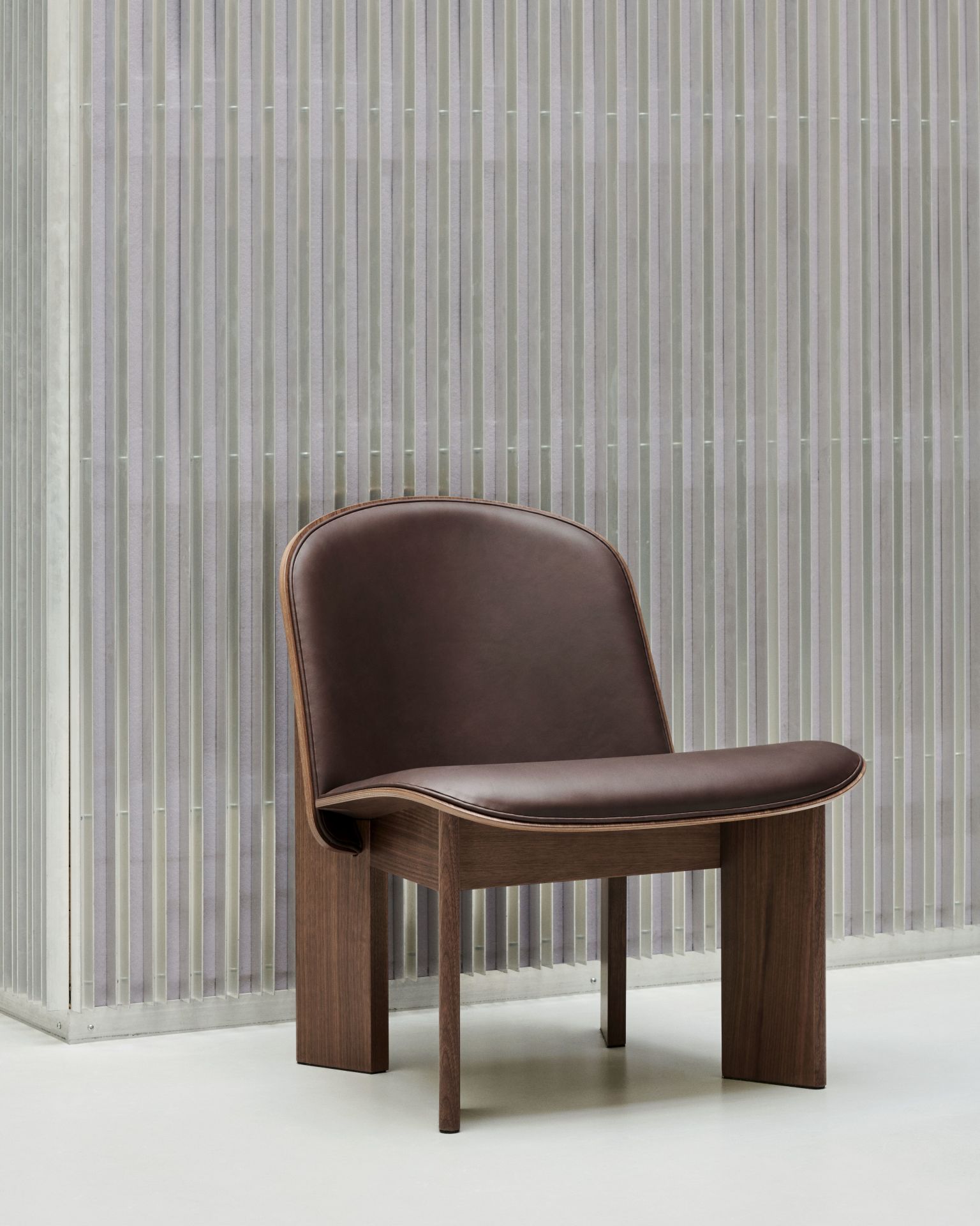Brauner Chisel Lounge Chair Sessel von Hay mit Vollpolsterung und Holzgestell vor gemusterter Wand.