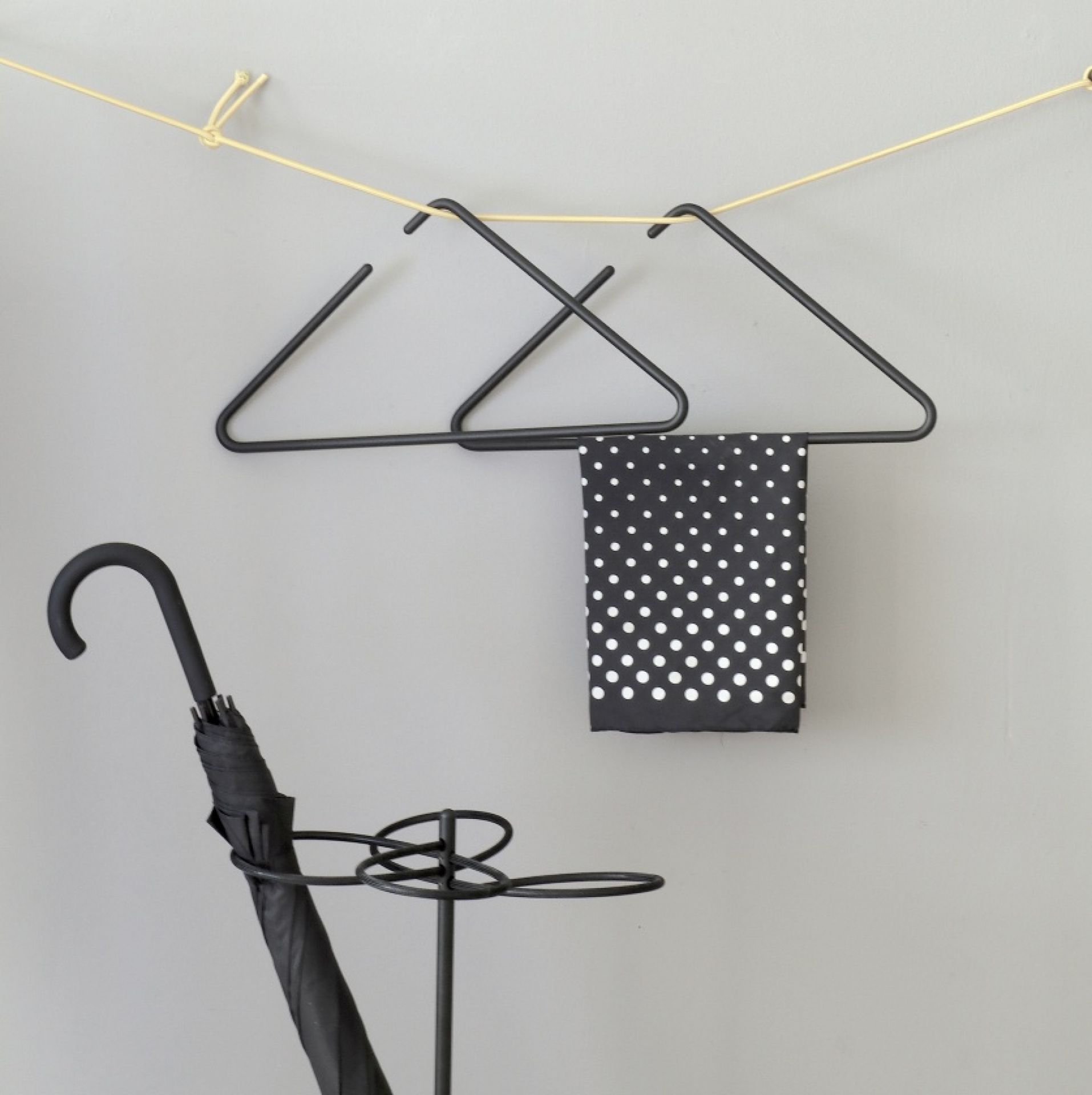 Roomsafari Triangle Kleiderbügel aus schwarzem Metall mit Schal und Schirmständer vor grauer Wand.