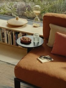 Orangefarbenes Sofa mit beigen und roten Vitra Kissen, Tischchen mit Snack, Buch, Brille und Sanduhr.