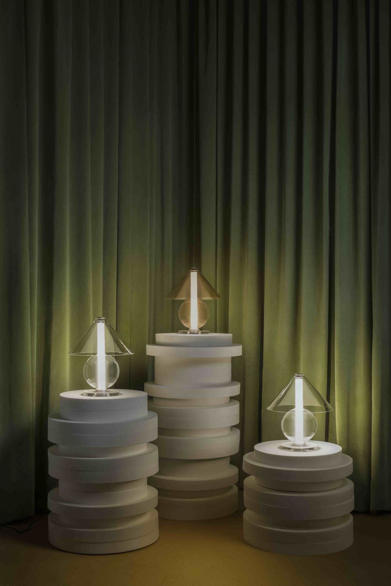 Drei Fragile Tischleuchten von Marset auf weißen Podesten vor grünem Vorhang.