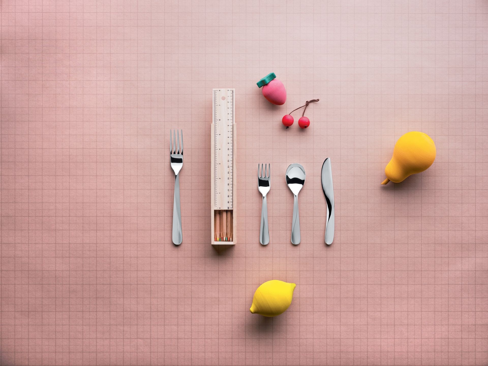 Alessi Kinderbesteck aus Edelstahl mit Messer, Gabel, Löffel, Stiften und Holzfrüchten auf kariertem Hintergrund.