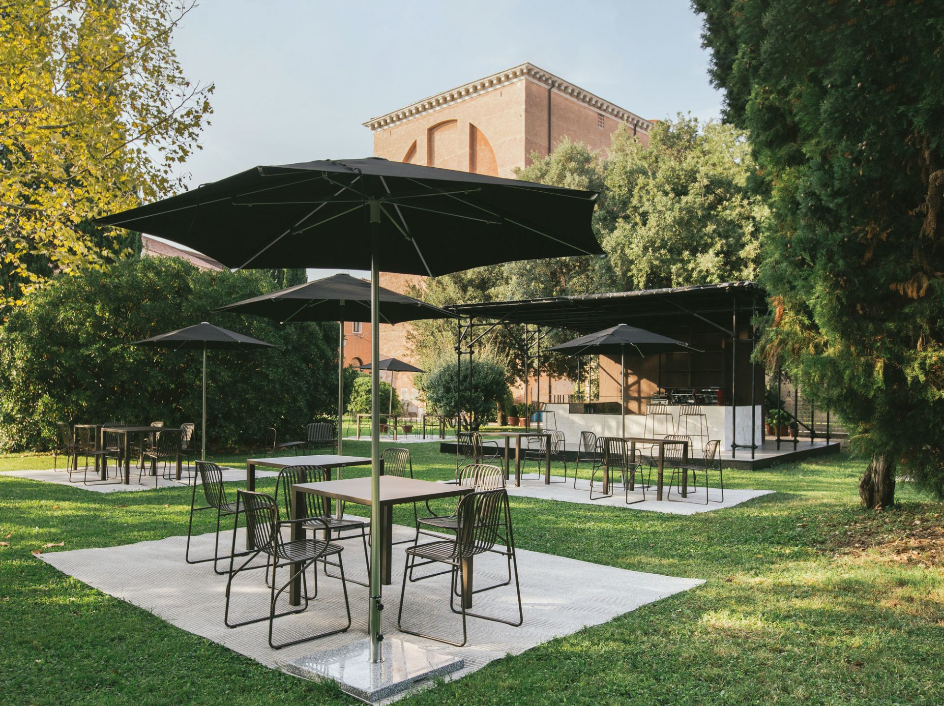 Riviera Armlehnstuhl von Emu: Schwarzer Metallstuhl im Außenbereich mit Tischen und Sonnenschirmen auf Rasen.