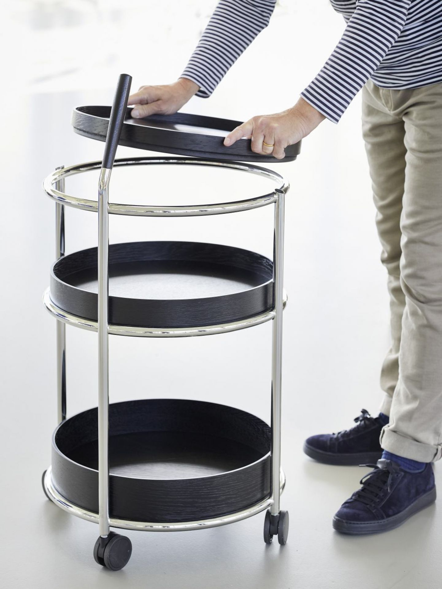 Tecta K6 Servierwagen mit Nussbaum-Ablagen und schwarzem Stahlrohr-Gestell. Person setzt oberes Tablett auf den Wagen.