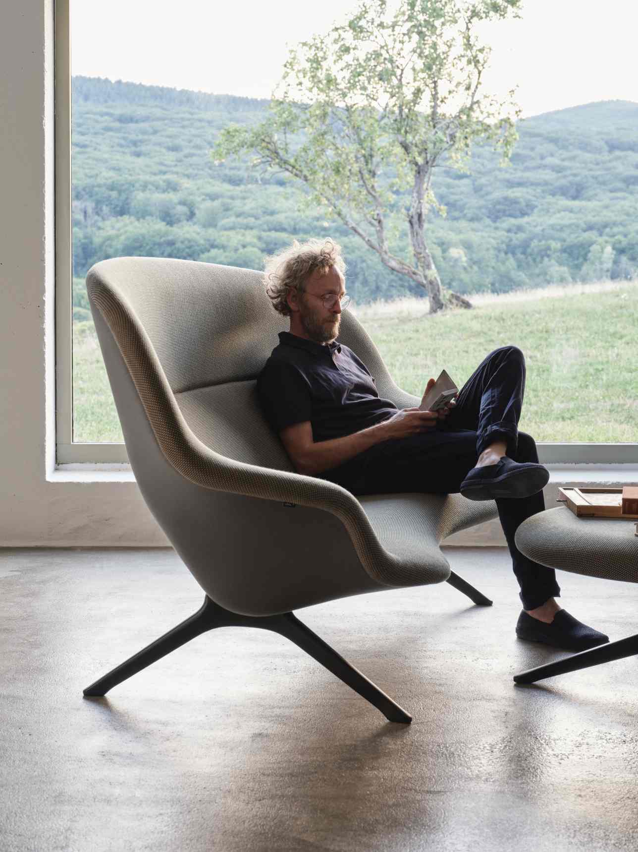 Mann liest in grauem Abalon Sessel mit Hocker von Vitra vor Fenster mit Landschaft.