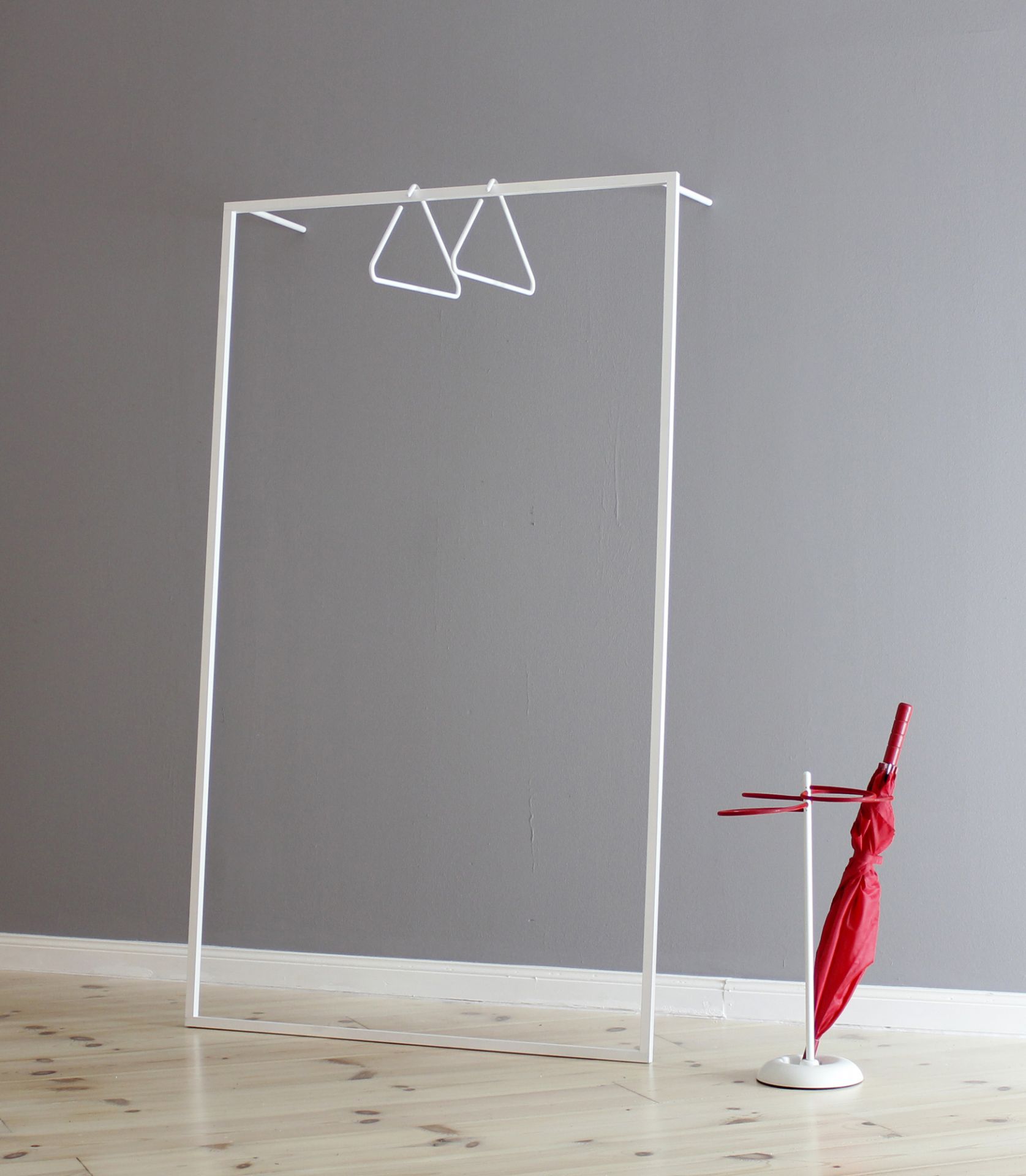 Weiße Leano Anlehngarderobe von Roomsafari mit zwei Kleiderbügeln und rotem Regenschirm vor grauer Wand.