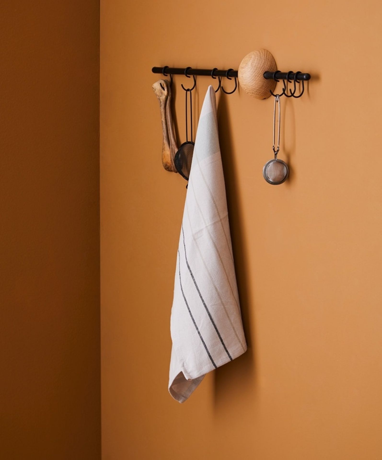 Schwarze Hoop Rack Wandgarderobe von Woud mit Küchentuch und Küchenutensilien an ockerfarbener Wand.