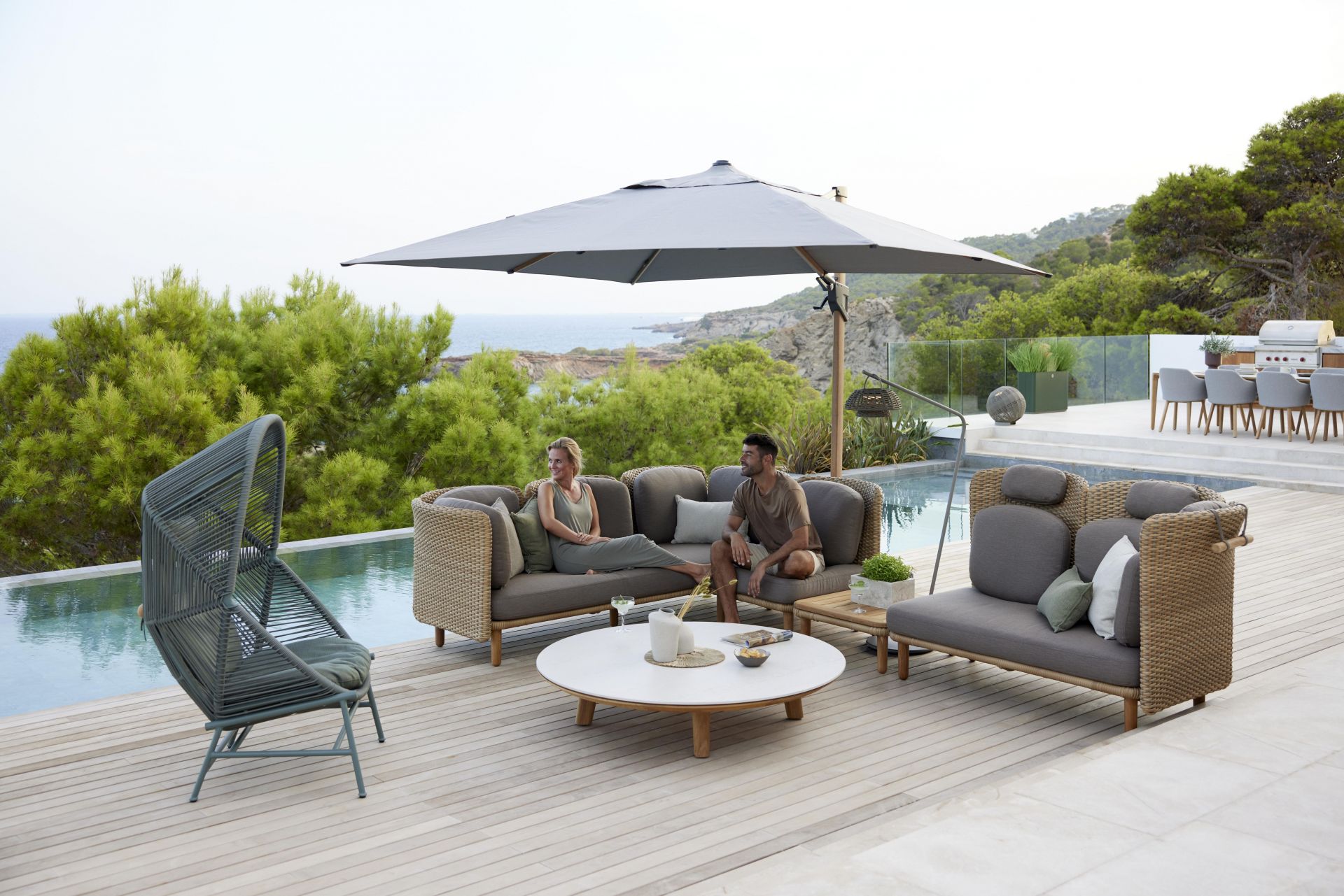 Arch Outdoor Couchtisch mit Teakplatte von Cane-Line auf einer Terrasse mit Pool und Sitzmöbeln.