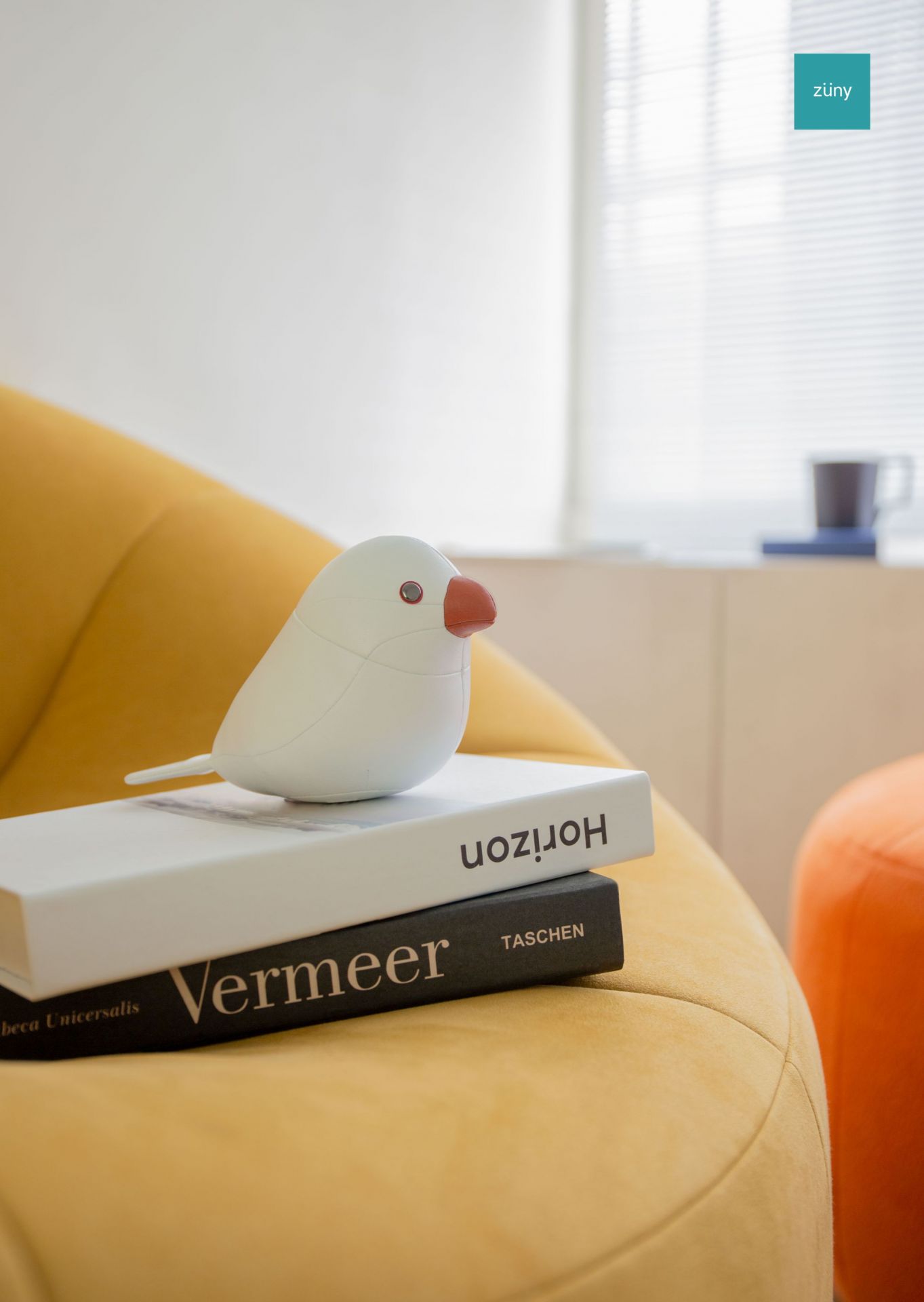 Weißer Java Spatz Briefbeschwerer von Züny auf zwei Büchern, sitzend auf einem gelben Sessel.