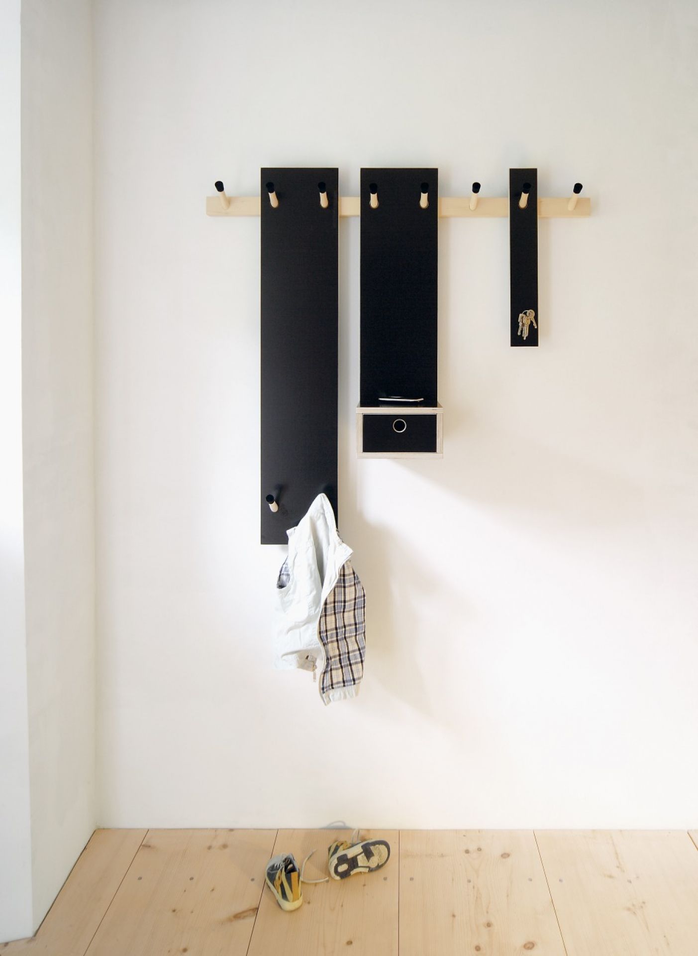 Schwarze Rechenbeispiel Kindergarderobe von Nils Holger Moormann an weißer Wand mit Jacke und Schuhen.