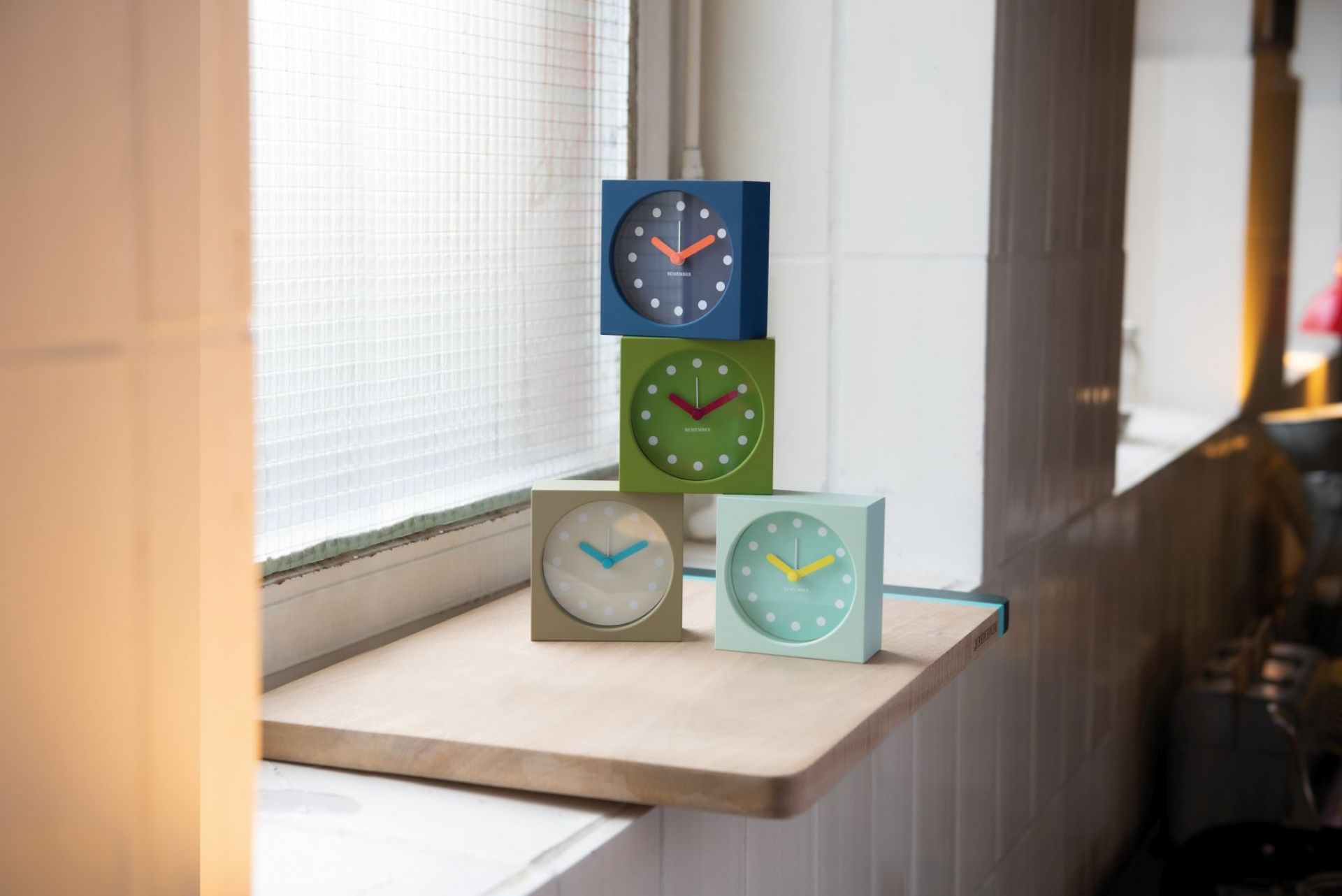 Stapel aus vier farbenfrohen Remember Tischuhren mit Wecker auf einem Holzbrett vor einem Fenster.