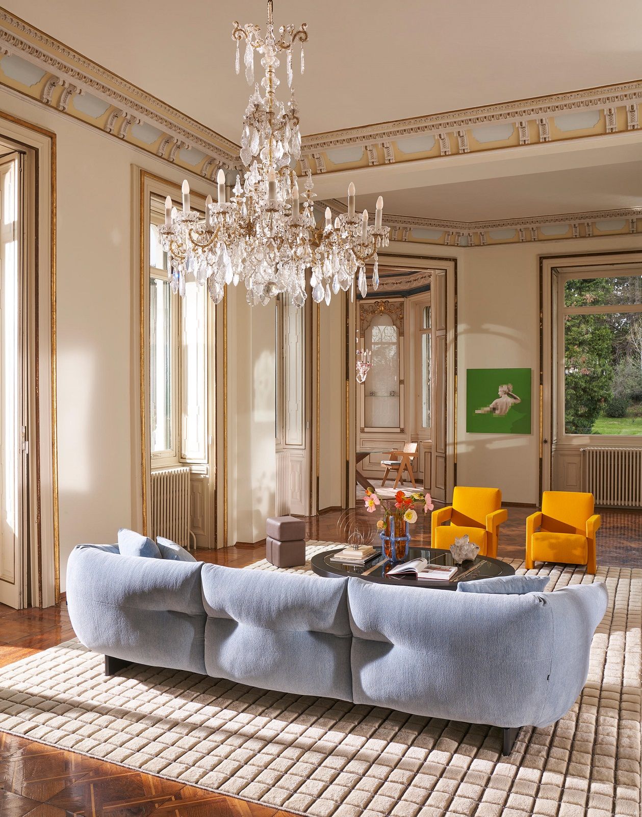 Helles Wohnzimmer mit blauem Moncloud Sofa von Cassina, Teppich, Kristalllüster und gelben Sesseln.