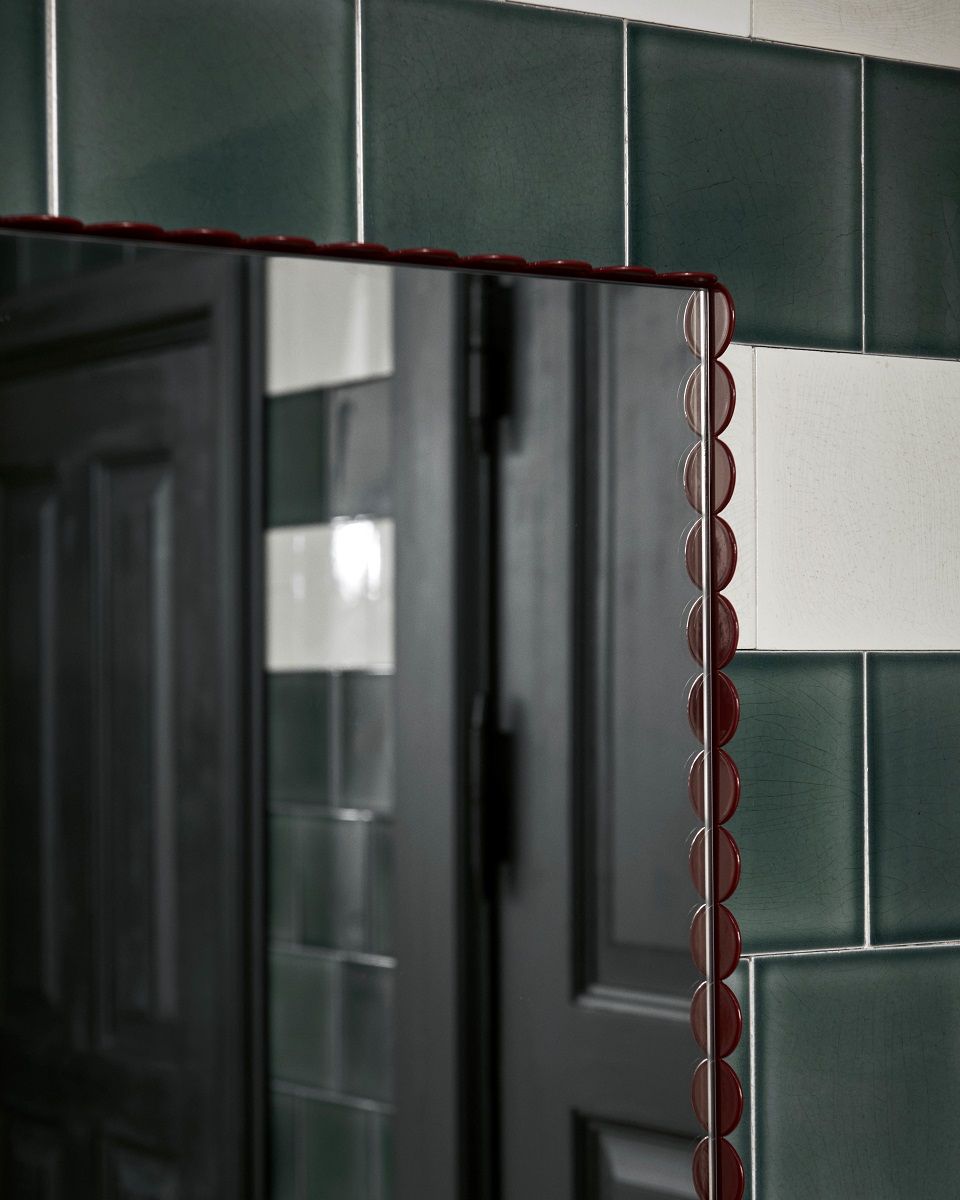 Detailaufnahme des Arcs Mirror Rectangle Spiegels von Hay mit burgunderrotem Rahmen vor grünen Fliesen.