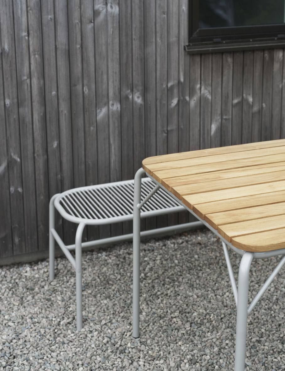 Normann Copenhagen Vig Bank Outdoor: Graue Gartenbank aus Metall und Teakholz Tischplatte auf Kies.