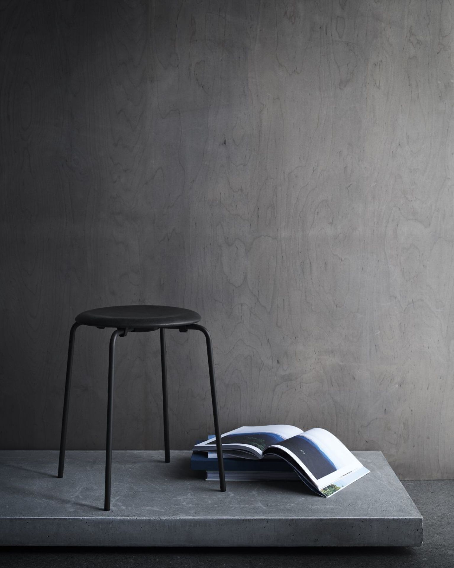 Schwarzer Dot Hocker von Fritz Hansen neben einem aufgeschlagenen Buch auf einer grauen Oberfläche.