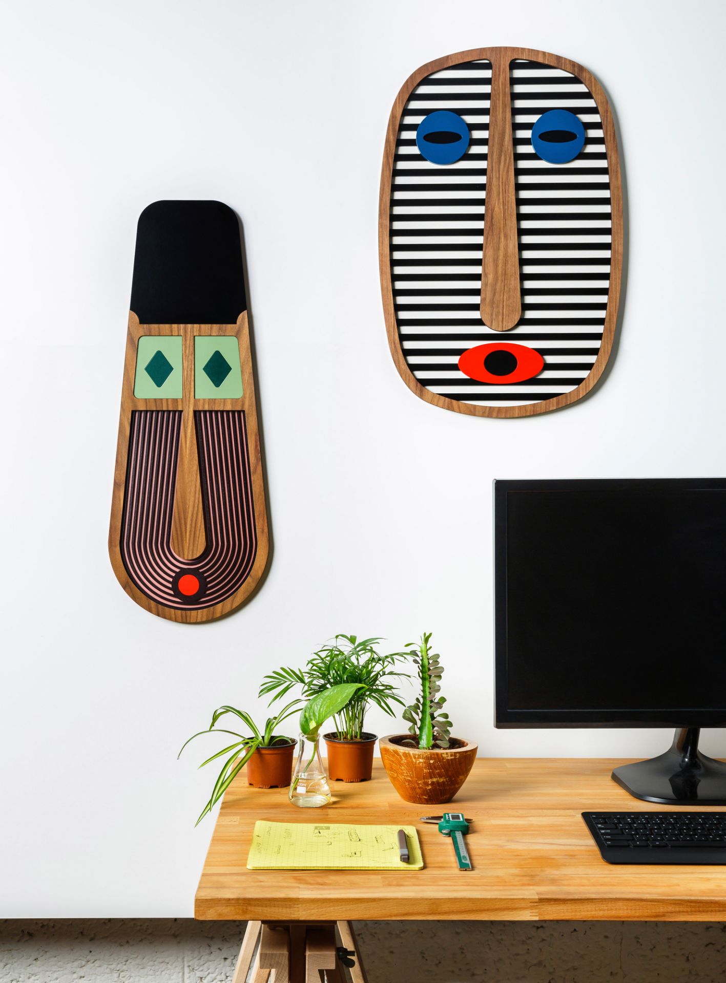 Zwei moderne afrikanische Umasqu Masken als Wandschmuck über einem Schreibtisch mit Pflanzen und Computer.