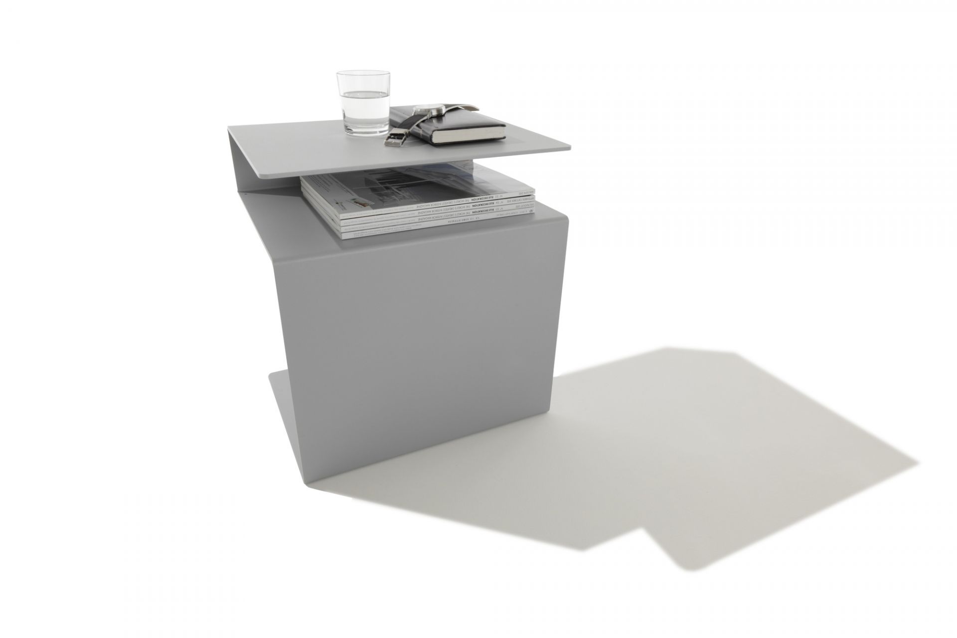 Grauer Huk Beistelltisch von Müller Möbelwerkstätten mit Glas, Notizbuch und Magazinen. Multifunktional und modern.