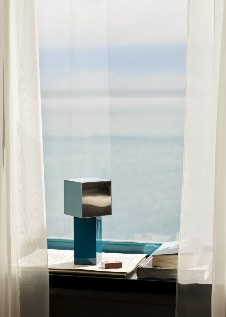 Pivot ATD7 Tischleuchte von AndTradition, blaue Akkuleuchte auf einem Tisch vor einem Fenster.