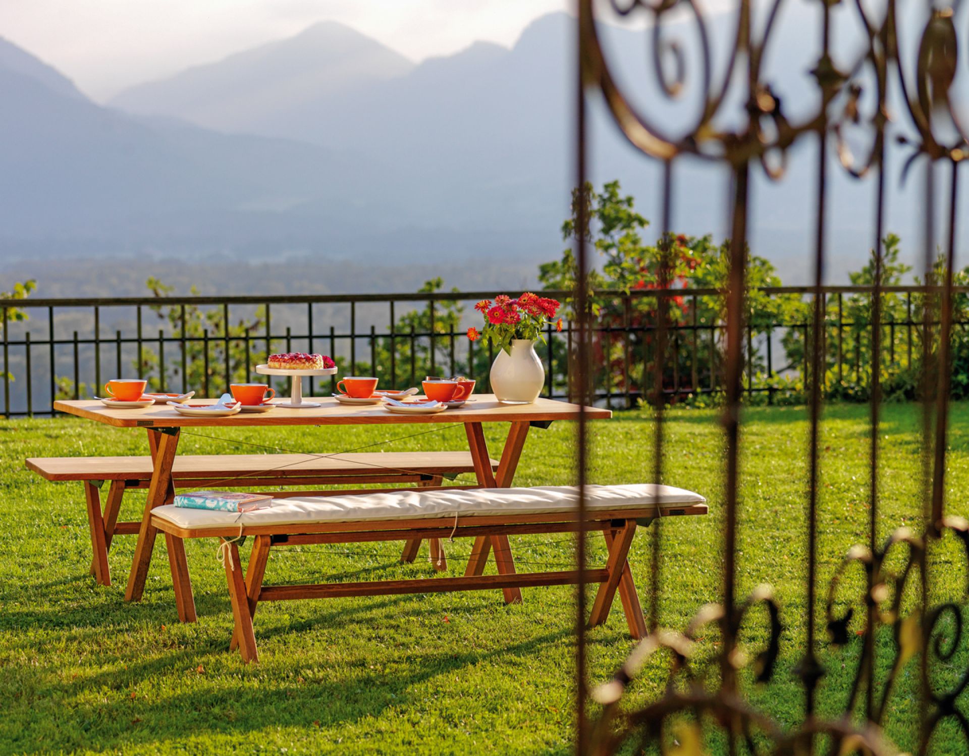Weishäupl Cross Bank klappbar mit Rückenlehne im Garten, gedeckter Tisch mit Gebäck und Getränken.