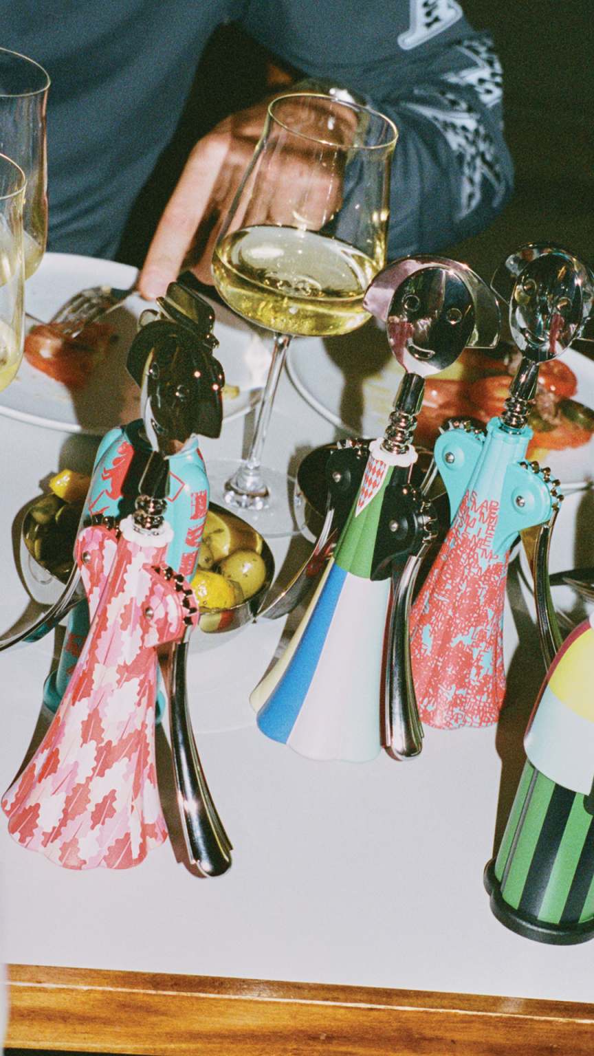 Mehrere bunte Alessi Anna G. Korkenzieher auf einem Tisch mit Essen und Weingläsern.