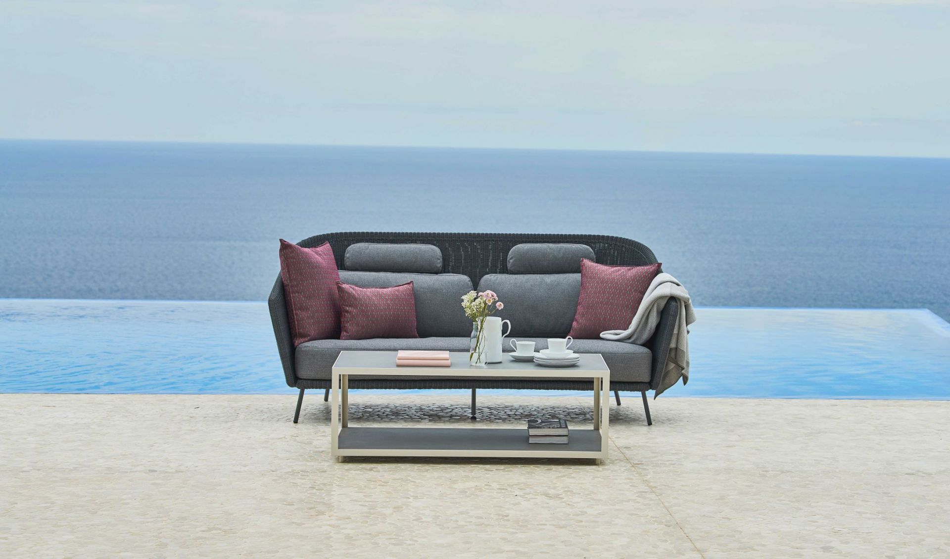 Mega Outdoor Sofa von Cane-Line mit Kissen und Tisch vor Pool und Meer.