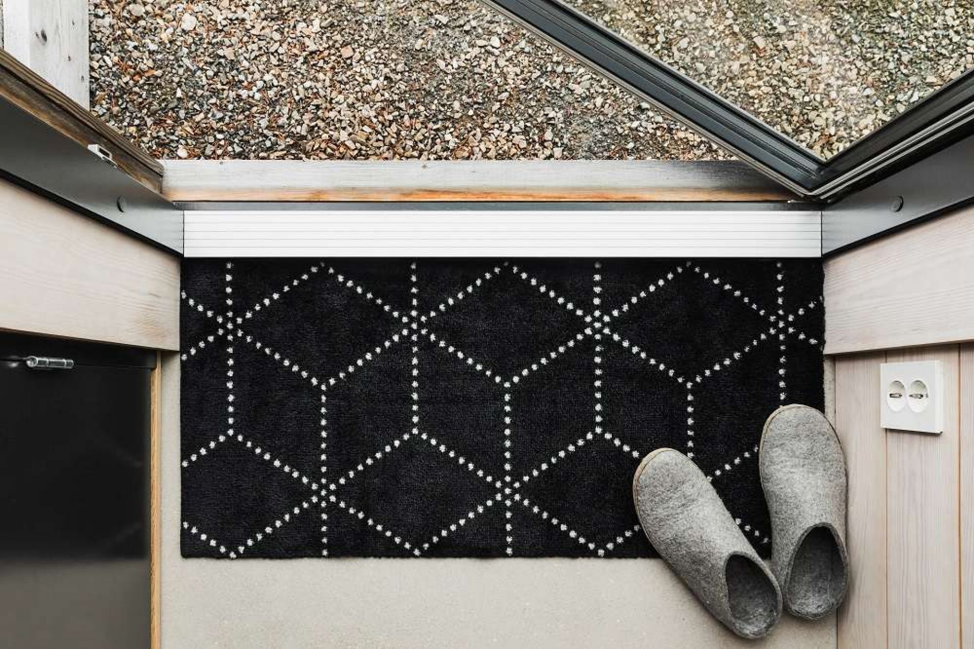 Schwarze Hagl Heymat Fußmatte mit geometrischem Muster vor einer Haustür, daneben graue Hausschuhe.