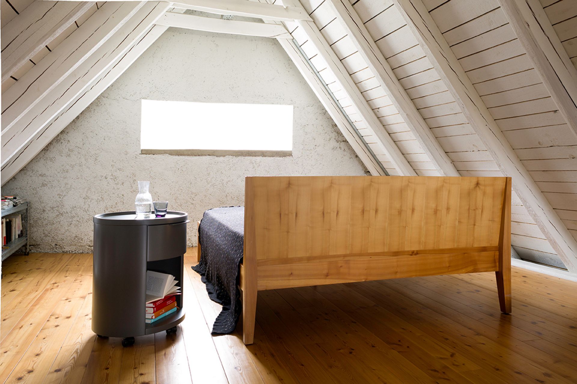 Conga Rundkommode von Schönbuch, grau, mit Rollen, neben einem Bett in einem Dachzimmer.