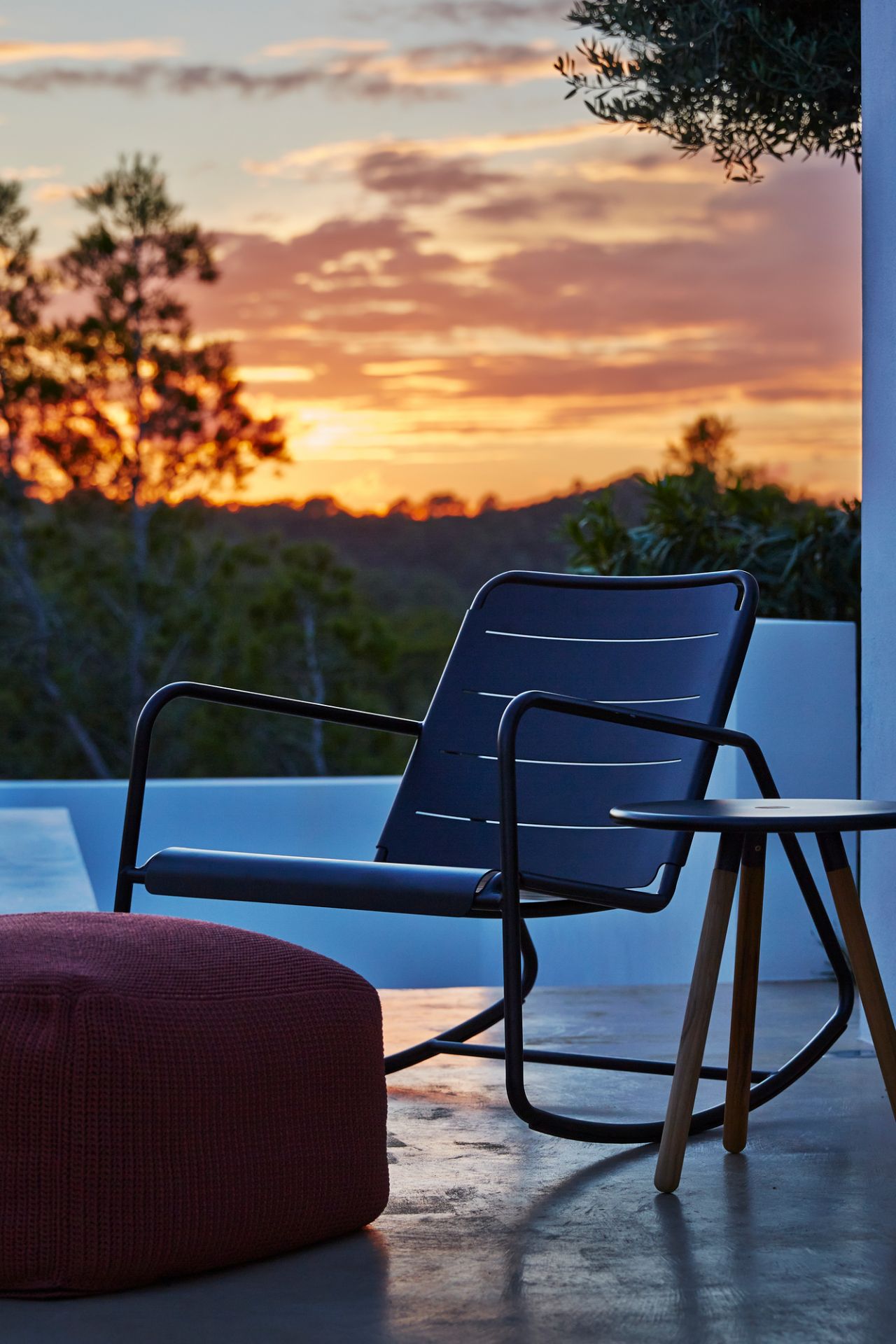 Cane-Line Schaukelstuhl für den Außenbereich, dunkelblau, mit Hocker und Beistelltisch bei Sonnenuntergang.