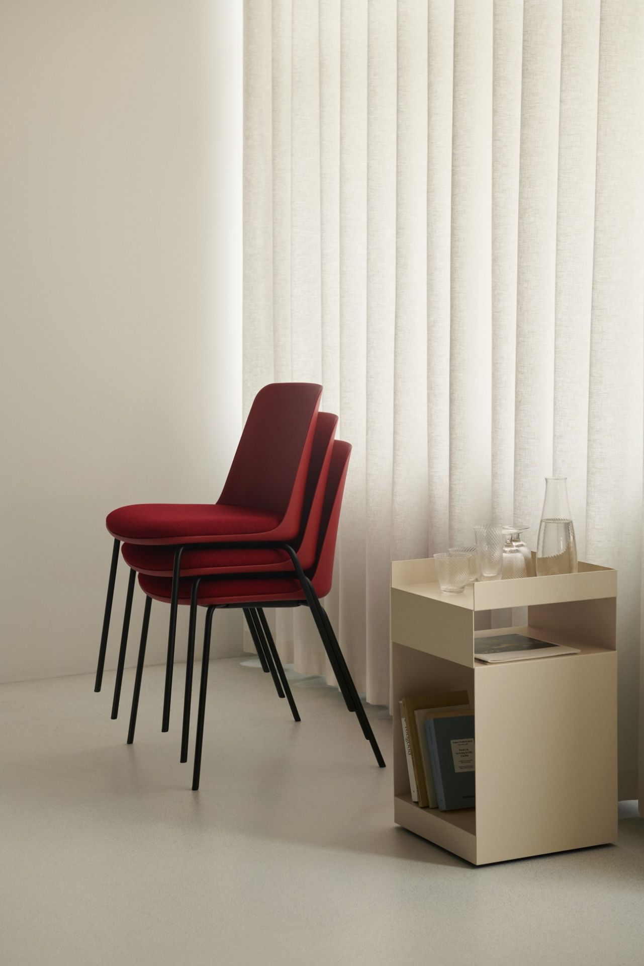Gestapelte rote AndTradition Esszimmerstühle mit schwarzen Beinen neben einem beigen Beistelltisch im minimalistischen Design.