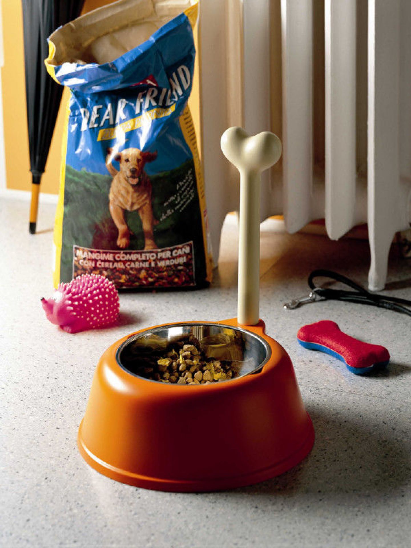 Oranger Alessi Lupita Hundenapf mit Knochen-Design, gefüllt mit Hundefutter, umgeben von Spielzeug.