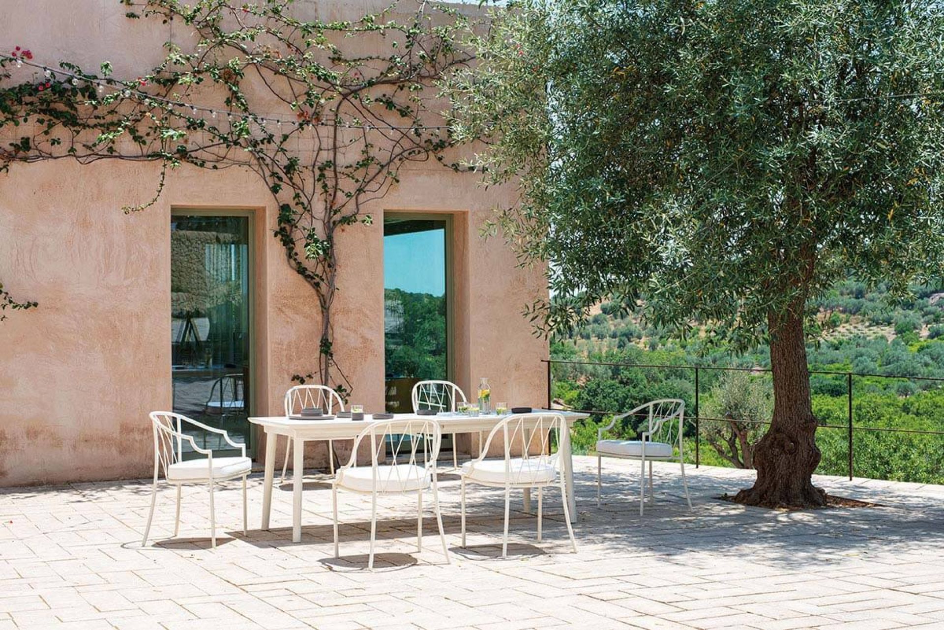 Emu Plus4 Ausziehtisch im Außenbereich mit weißen Stühlen auf einer Terrasse unter einem Baum.