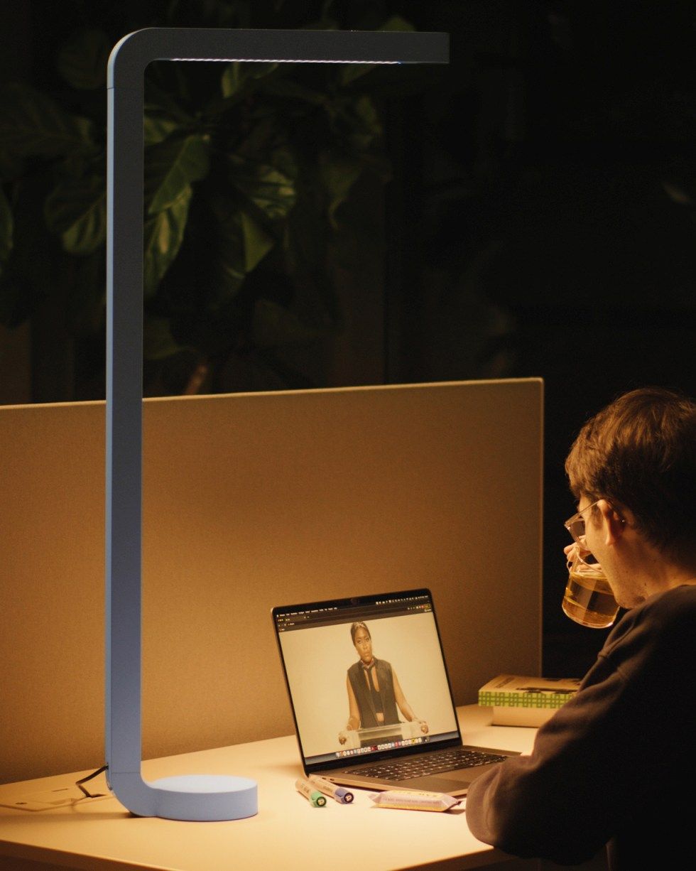 Graue Team Home Tischleuchte beleuchtet Schreibtisch mit Laptop und Person beim Trinken, ideal für Homeoffice.